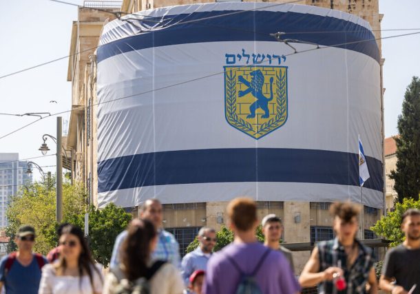 La gente pasa frente a una bandera gigante del municipio de Jerusalén colgada frente a la Ciudad Vieja el 26 de mayo de 2022, antes del Día de Jerusalén. (Yonatán Sindel/Flash90)
