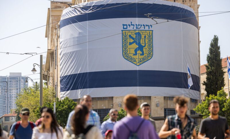 La gente pasa frente a una bandera gigante del municipio de Jerusalén colgada frente a la Ciudad Vieja el 26 de mayo de 2022, antes del Día de Jerusalén. (Yonatán Sindel/Flash90)